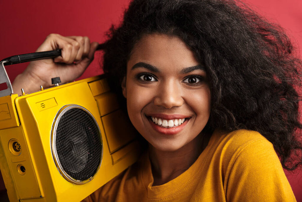 Image of beautiful brunette african american woman with curly hair laughing and holding vintage boombox isolated over red background - Fotografie, Obrázek