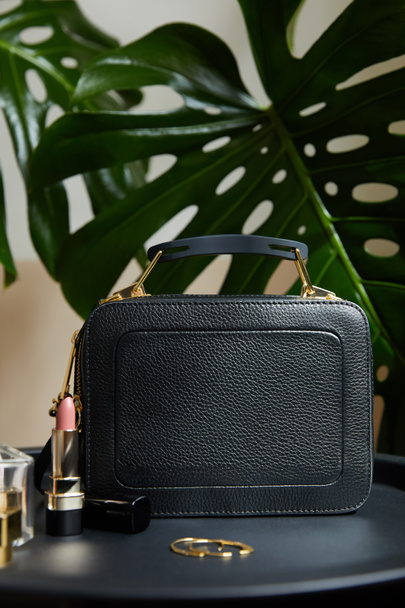 leather handbag near golden earrings and lipstick on black table near tropical leaves - Fotoğraf, Görsel