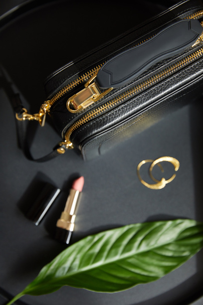top view of leather handbag near golden earrings and lipstick on black table with green leaf - Foto, Imagem