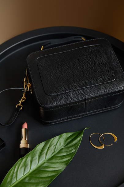 leather handbag near golden earrings and pink lipstick on black table with tropical leaf - Fotoğraf, Görsel