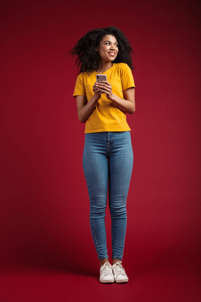 Full length image of beautiful brunette african american woman with curly hair smiling and holding cellphone isolated over red background - Foto, Imagen