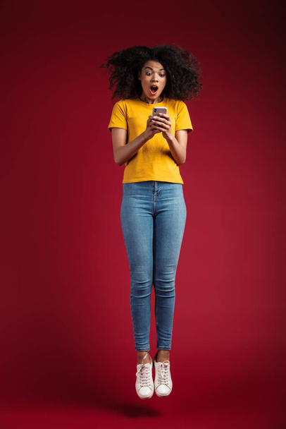 Ganzes Bild der schönen brünetten afrikanisch-amerikanische Frau mit lockigem Haar jubelt und hält Handy isoliert über rotem Hintergrund - Foto, Bild