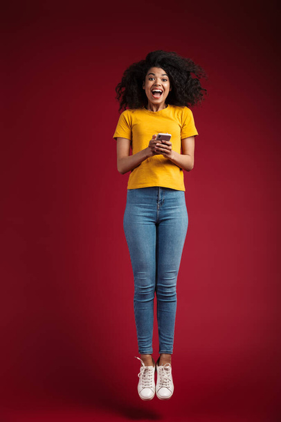 Full length image of beautiful brunette african american woman with curly hair smiling and holding cellphone isolated over red background - Φωτογραφία, εικόνα