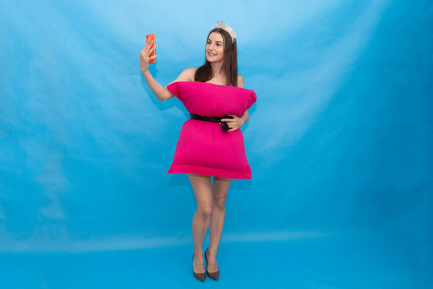 счастливая женщина в розовом платье подушки сделать селфи фото на мобильном телефоне для сумасшедшего вызова подушки на синем фоне из-за того, что в доме изолированы. Модная девушка. Подушка по дому
 - Фото, изображение