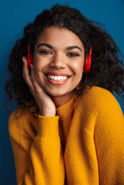 Image of a pretty young smiling african woman posing isolated over blue wall background listening music with headphones. - Valokuva, kuva