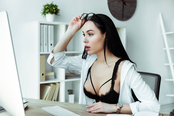 Excited secretary in bra holding eyeglasses and looking at computer monitor in office  - Фото, изображение
