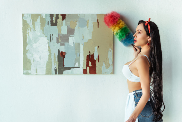 Vue latérale de fille sexy en soutien-gorge et tablier nettoyage peinture avec brosse à poussière à la maison
  - Photo, image