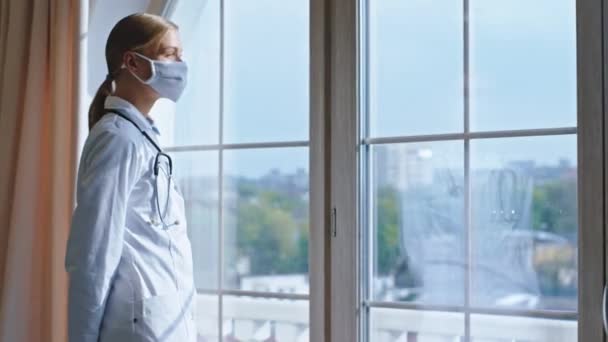 Young woman doctor with a protective mask in her face looking through the panoramic window in the hospital she take a break have a tired face Covid-19 concept - Video