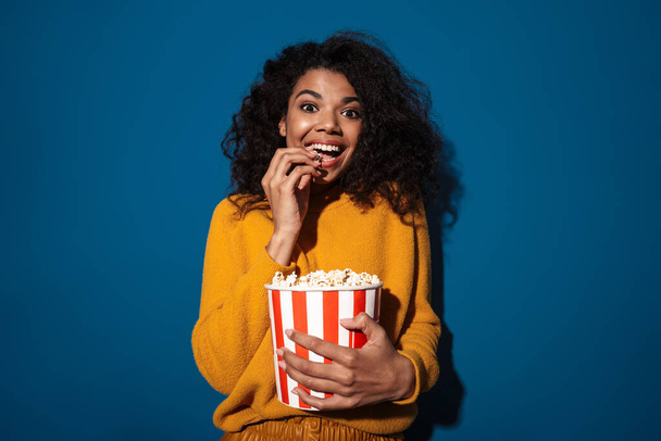 Photo of positive young african woman posing isolated over blue wall background holding popcorn watch film. - Foto, afbeelding