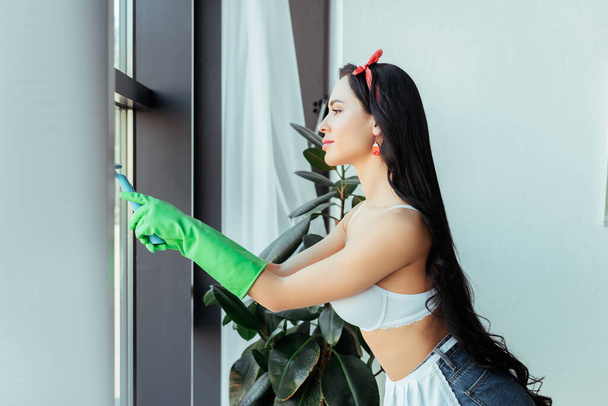 Side view of sensual woman in rubber gloves cleaning window at home  - Фото, зображення