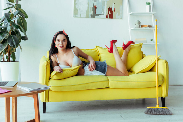 Sensual smiling woman looking at camera while lying on couch near broom  - Photo, image