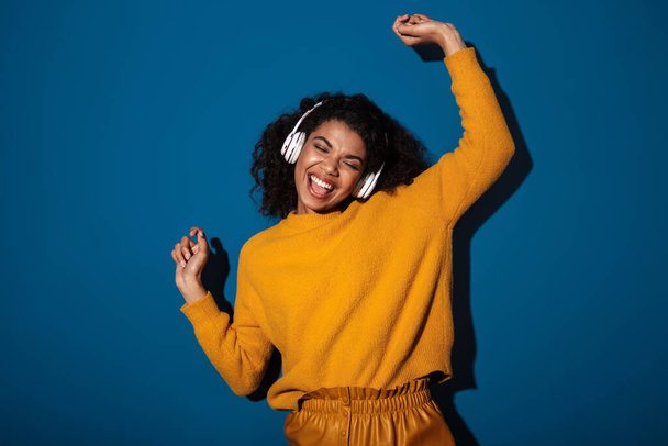 Photo of happy positive young african woman posing isolated over blue wall background listening music with headphones. - Zdjęcie, obraz