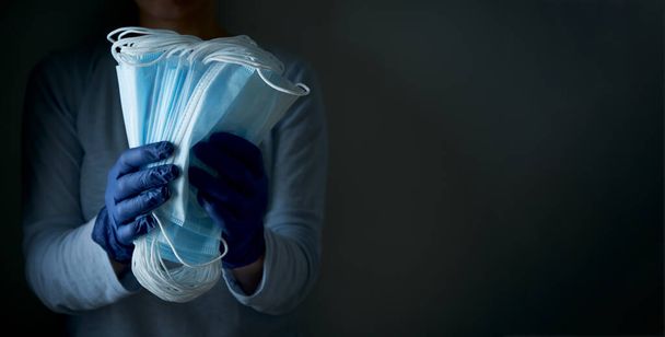 A woman holds a stack of face protective medical masks in latex disposable gloves on a dark background, close-up. Save lives. - Foto, Imagen