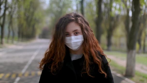 Nuori tyttö lääketieteellisessä naamiossa seisoo ulkona puistossa. COVID-19:n käsite
 - Materiaali, video