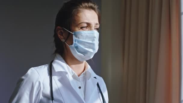 Modern hospital in the stuff room portrait of a tired and sad woman doctor wearing a protective mask she looking through the window Coronavirus pandemic 2019 - Video