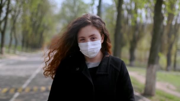 Een jong meisje met een medisch masker staat buiten in een park. Het concept van COVID-19 - Video