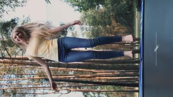 トランポリンでジャンプする幸せな女性。美しい若いですブロンド裸足の女性で光夏のシャツとジーンズ見てカメラと笑顔ジャンプで大トランポリンで背の高い木の周り. - 映像、動画