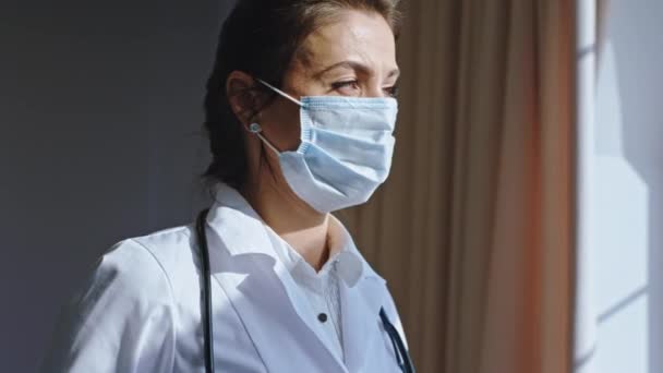 Mature beautiful woman with a protective mask take a break in her office room she looking through the hospital window with a panoramic view - Filmmaterial, Video