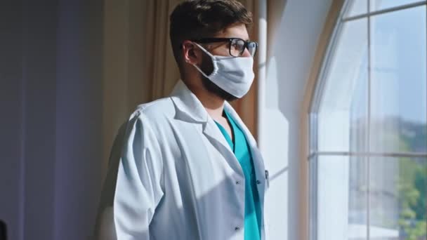 In a modern hospital young man doctor with a protective mask have a little break he take a look though the panoramic window and have a thinking face. Shot on ARRI Alexa Mini - Video