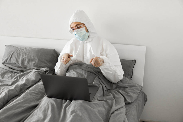 Man in protective white suit and medical mask is working from home in a bed with laptop and speaking with his friend or business partners via webcam. Coronavirus epidemic. Remote work during pandemic. - Photo, image