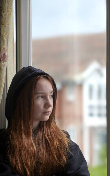 Αναστατωμένη έφηβη με μαύρη κουκούλα με κόκκινα μαλλιά να στέκονται δίπλα στο παράθυρο και να κοιτάζουν έξω από το παράθυρο στο σπίτι κατά τη διάρκεια της απομόνωσης - Φωτογραφία, εικόνα