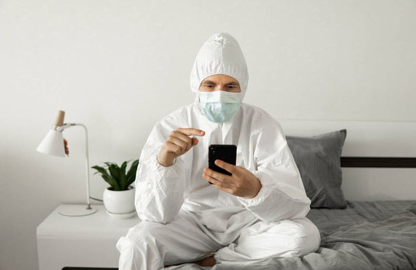 Man in protective white suit and medical mask is using a phone at his home sitting on a bed because of coronavirus epidemic. Remote work during pandemic. Stay home during COVID-19 quarantine concept. - Photo, image