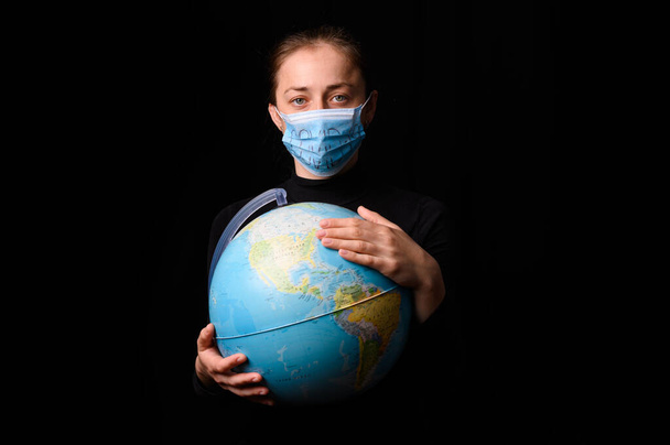 Woman hands holding the Earh globe in the hands. Woman in the doctor breath medicine mask. Coronavirus protection for people all continents and the world. Isolated on a black background. Concept stop covid-19. - Фото, изображение
