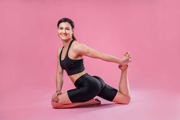 Entraîneur de fitness fille sportive vêtu d'un haut noir et leggings noirs assis sur le sol sur un fond rose dans le studio. Exercice d'étirement musculaire des jambes et sourire
. - Photo, image