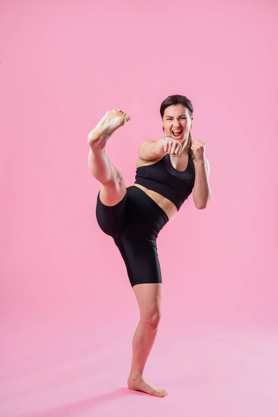 Slim woman is kicking the air, with hand and leg up . Athletic girl doing boxing exercises, making a direct kick. Photo of a young girl isolated on a pink background. Strength and motivation. - Foto, Bild