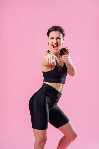 Slim woman is kicking the air, with hand and leg up . Athletic girl doing boxing exercises, making a direct kick. Photo of a young girl isolated on a pink background. Strength and motivation. - 写真・画像