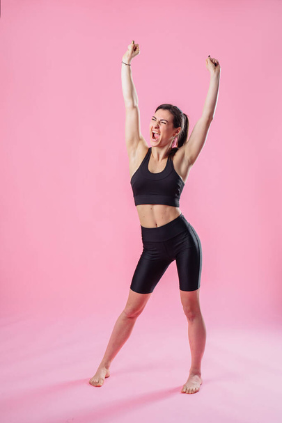 Full-length portrait. Attractive girl fitness trainer joyfully stretching. Woman with a beautiful body raised her hands up. Isolated on a pink background in a photo studio. - Photo, Image