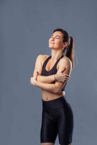 Pretty Hispanic personal trainer and diet coach standing isolated on gray background. Health and fitness concept - portrait of an caucasian american girl posing with fitness clothes. Hugging herself - Foto, afbeelding