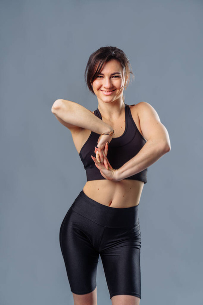 A beautiful sports girl in black sportswear is doing a warm-up before practicing sports at the studio. The concept of a healthy lifestyle. Athletic woman stretches her arms before gym workout. - Foto, imagen
