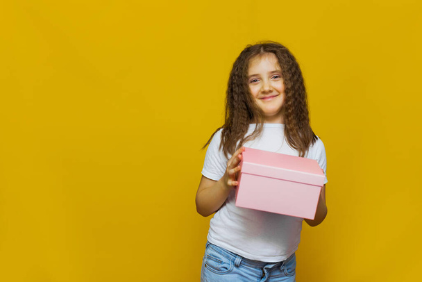 Little cute smiling girl in white T-shirt holding pink gift box on Yellow background. child holding gift box. - Foto, Bild