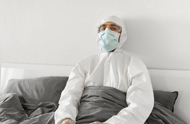 Portrait of man in protective white suit and face mask sitting in his bed in his bedroom because of covid-2019 and wait for a end of coronavirus quarantine. Self isolation. - Foto, imagen