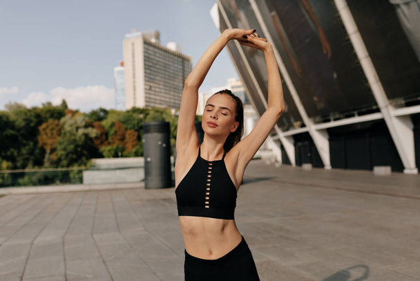 Lovely charming woman in sport trendy uniform stretching before training outdoor on sport area in summer sunny day. Confident and fit female preparing to training closing eyes and smiling.  - Photo, image