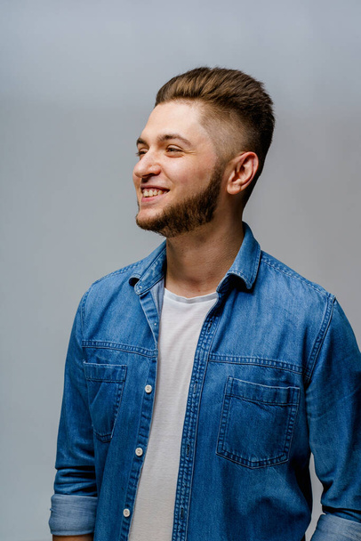 Young handsome man stands over white background and touches smiles after visiting barbershop. Confident man wears casual jeans shirt and looks in camera. Bearded caucasian business man. - Foto, Imagem