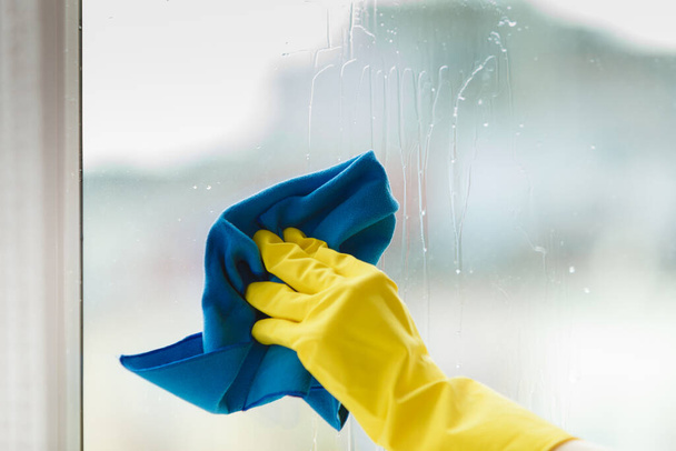 Female hand in yellow gloves cleaning window with blue rag and spray detergent. Spring cleanup, housework concept - Foto, Bild