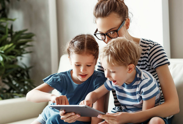 Vidám fiatal anya kisfiúval és kislánnyal ül a kanapén, és videojátékot játszik tableten, miközben együtt töltik az időt a Hom-ban. - Fotó, kép