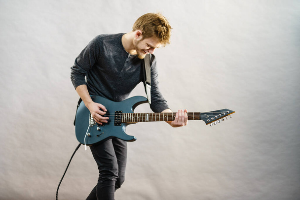 Young bearded man with electric guitar. Adult person is holding instrument and playing. Hobby, music concept, on grey - Foto, afbeelding