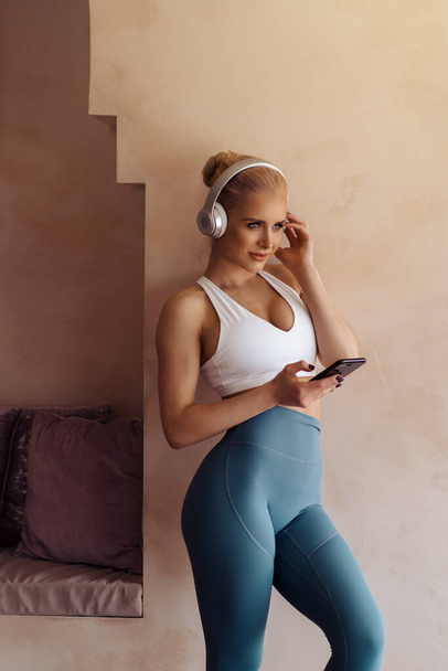 Beautiful healthy fit beautiful young woman in headphones holding smartphone in hands. Attractive sporty woman listening to the music while doing home workout. - 写真・画像