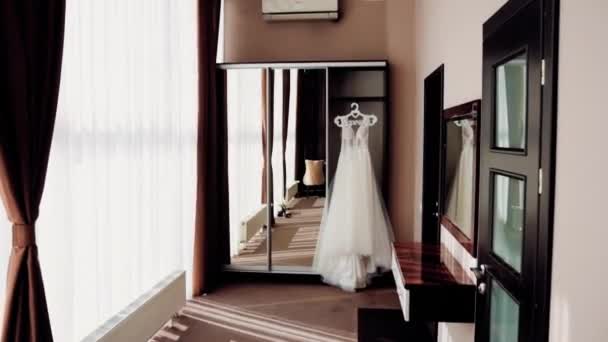 In een mooie bruine hotelkamer aan een grote gespiegelde kastdeur hangt een lange witte jurk aan het raam - Video
