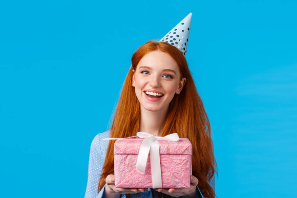 Hediye zamanı. Neşeli kızıl saçlı genç, kolejli kız doğum gününü kutluyor, elinde sevimli pembe bir hediye tutuyor ve doğum günü şapkası takıyor, neşeyle gülümsüyor mavi arka planda. - Fotoğraf, Görsel