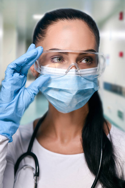 Girl in protective glasses, medical mask and blue gloves on blured hospital background . close-up portrait of a woman in a transparent mask. way to protect against coronavirus. Covid-2019, Pandemic 2020. Soft focus - Photo, image