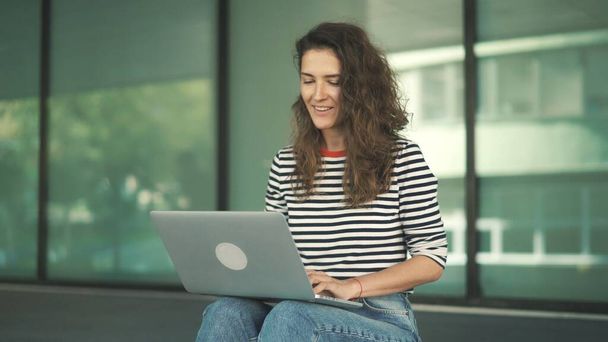 Attraktive junge Frau mit langen lockigen Haaren und gestreiftem Hemd sitzt auf der Treppe eines Gebäudes in der Stadt und arbeitet am Laptop - Foto, Bild