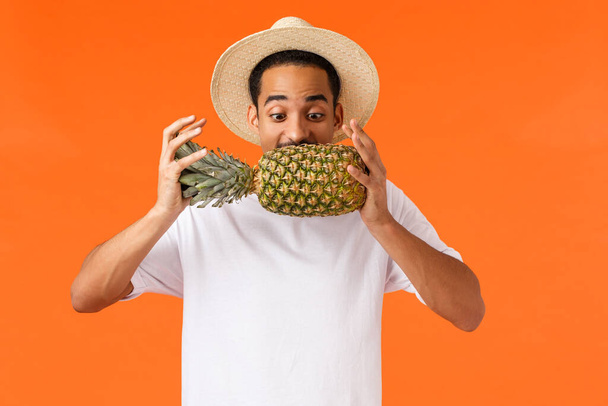 Funny handsome hipster guy, african american tourist enjoying vacation, trying bite pineapple, looking at fruit, standing orange background joyful, like travel hot countries during winter season - Fotografie, Obrázek