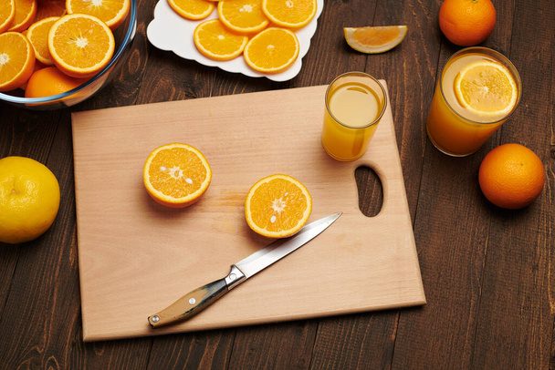 Fresh orange fruit whole and sliced on a wooden table, cutting board and kitchen knife. A plate full of citrus slices - natural and healthy food. Glass of fruit cocktail. - Photo, Image