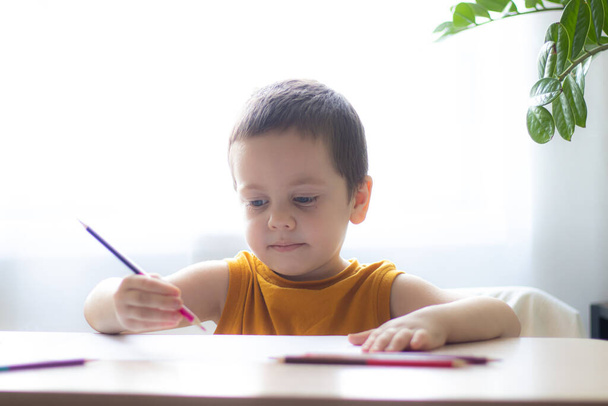 El niño dibuja con lápices en la mesa. La tarea. Un chico en la ventana con una flor. Aprendizaje online. Aislamiento de cuarentena. Estamos sentados en casa. Entretenimiento infantil con autoaislamiento. Ocio casero. Hobbies están dibujando
. - Foto, Imagen