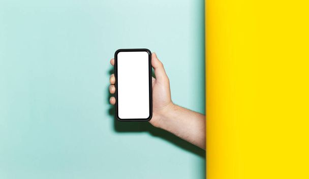 Close-up of male hand holding smartphone with mockup on two studio backgrounds of yellow and aqua menthe colors.  - Photo, Image