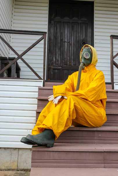 person in a gas mask and protective yellow costume sitting on the porch of the house. - Photo, Image